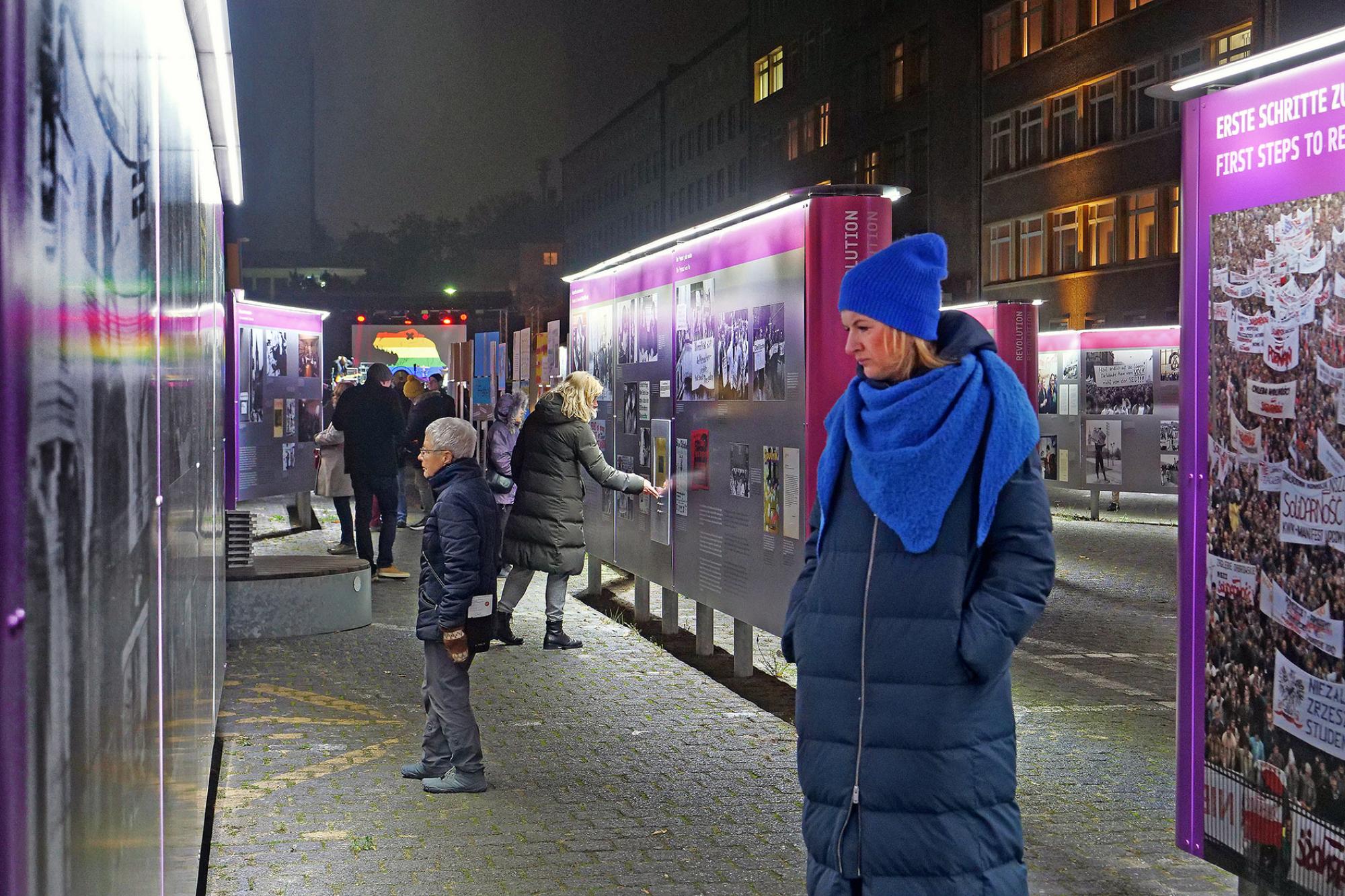 Menschen versammeln sich bei einer Open-Air-Veranstaltung auf dem Gelände der ehemaligen Stasi-Zentrale in Berlin; im Hintergrund das historische Gebäude, im Vordergrund bunte Infostände und Sitzgelegenheiten.