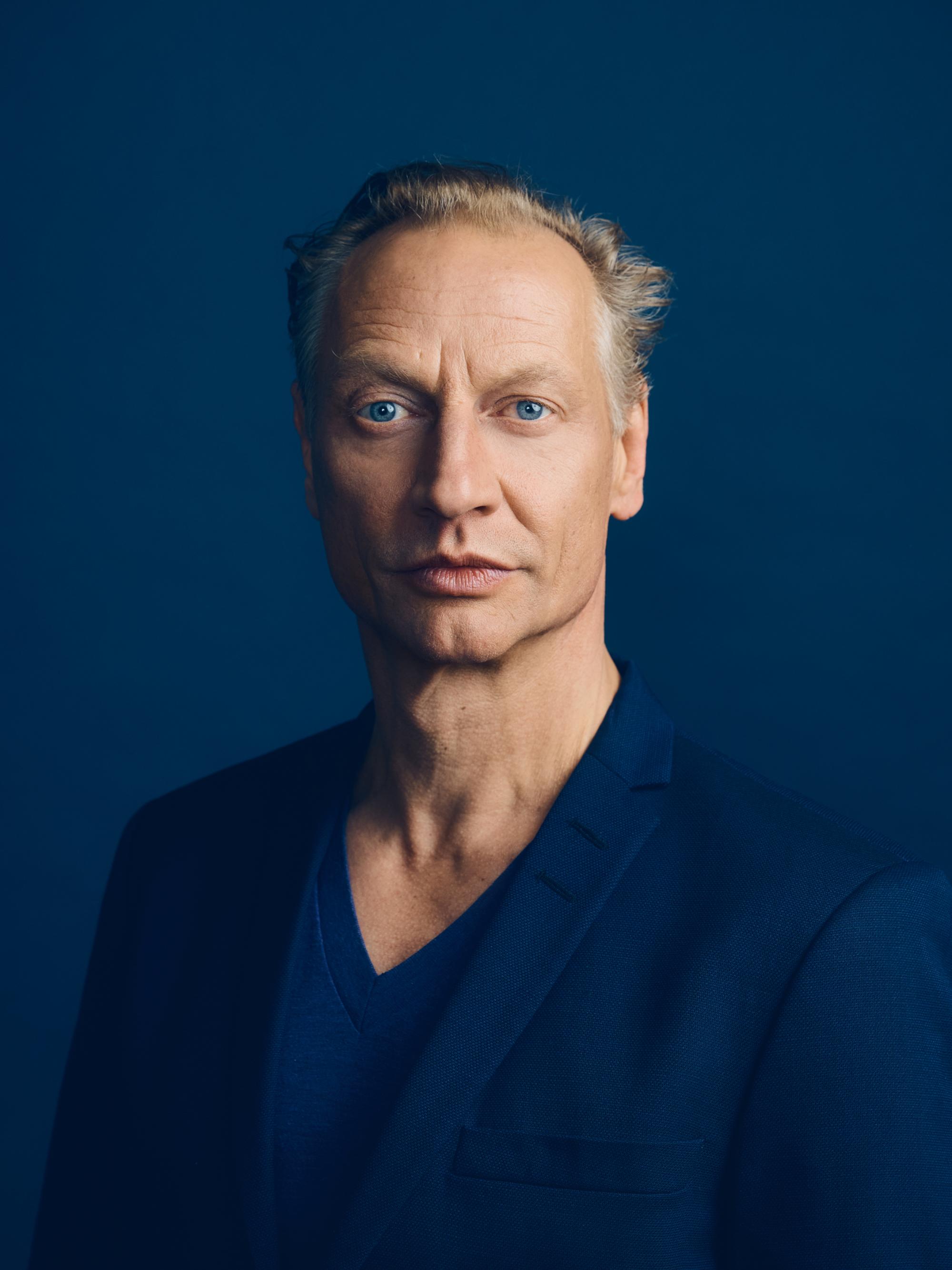 Porträt von Victor Schefé vor dunkelblauem Hintergrund, mit ernstem Gesichtsausdruck und blauen Augen, gekleidet in ein dunkelblaues Sakko und Shirt.