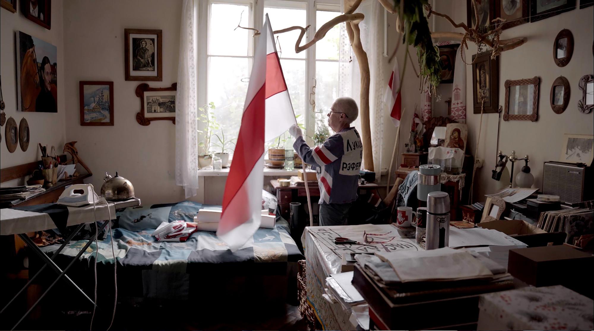 Ältere Frau in einem mit Bildern und Erinnerungsstücken dekorierten Wohnzimmer hält eine weiß-rot-weiße belarussische Flagge am Fenster.