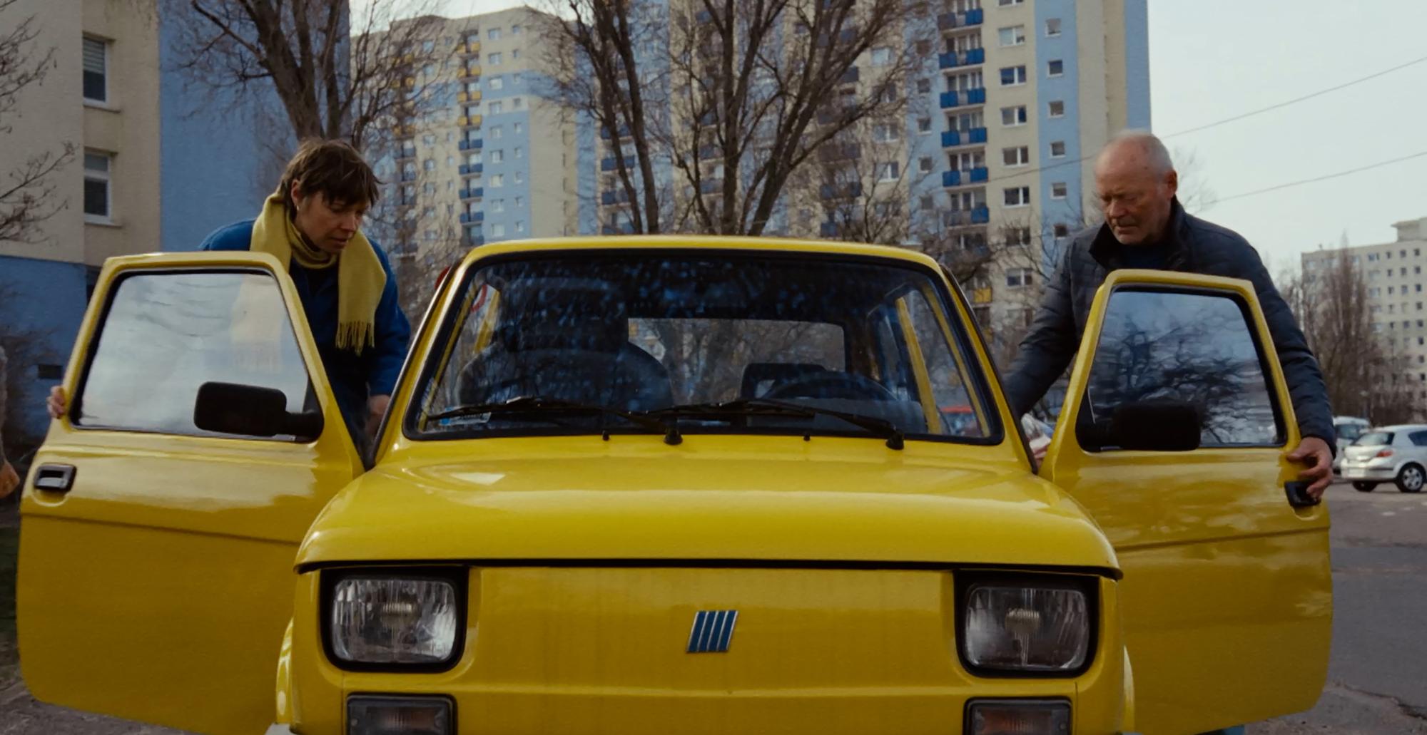 Ein Filmstill aus dem Film "Die noch unbekannten Tage": Zwei Menschen steigen in ein gelbes Auto aus DDR-Zeiten ein.