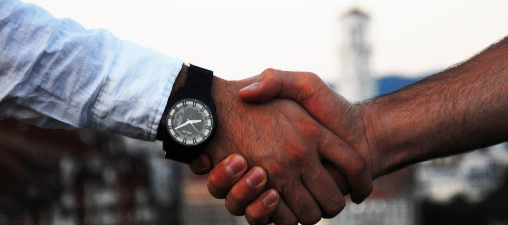 Ein Händedruck zwischen zwei weißen Männern. Ein Arm trägt eine schwarze Uhr und ein blaues Hemd.