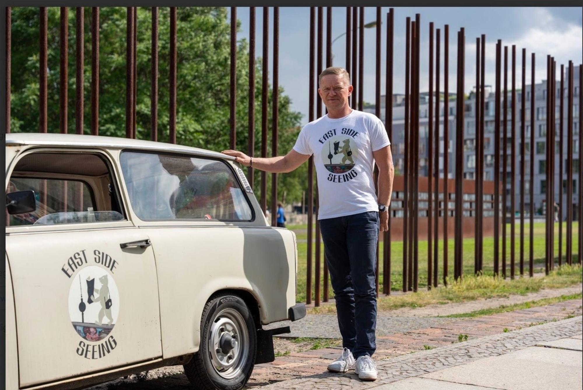 Ein Foto von Markus Wiehler vor der Gedenkstätte Berliner Mauer, mit einem alten Trabi-Auto.