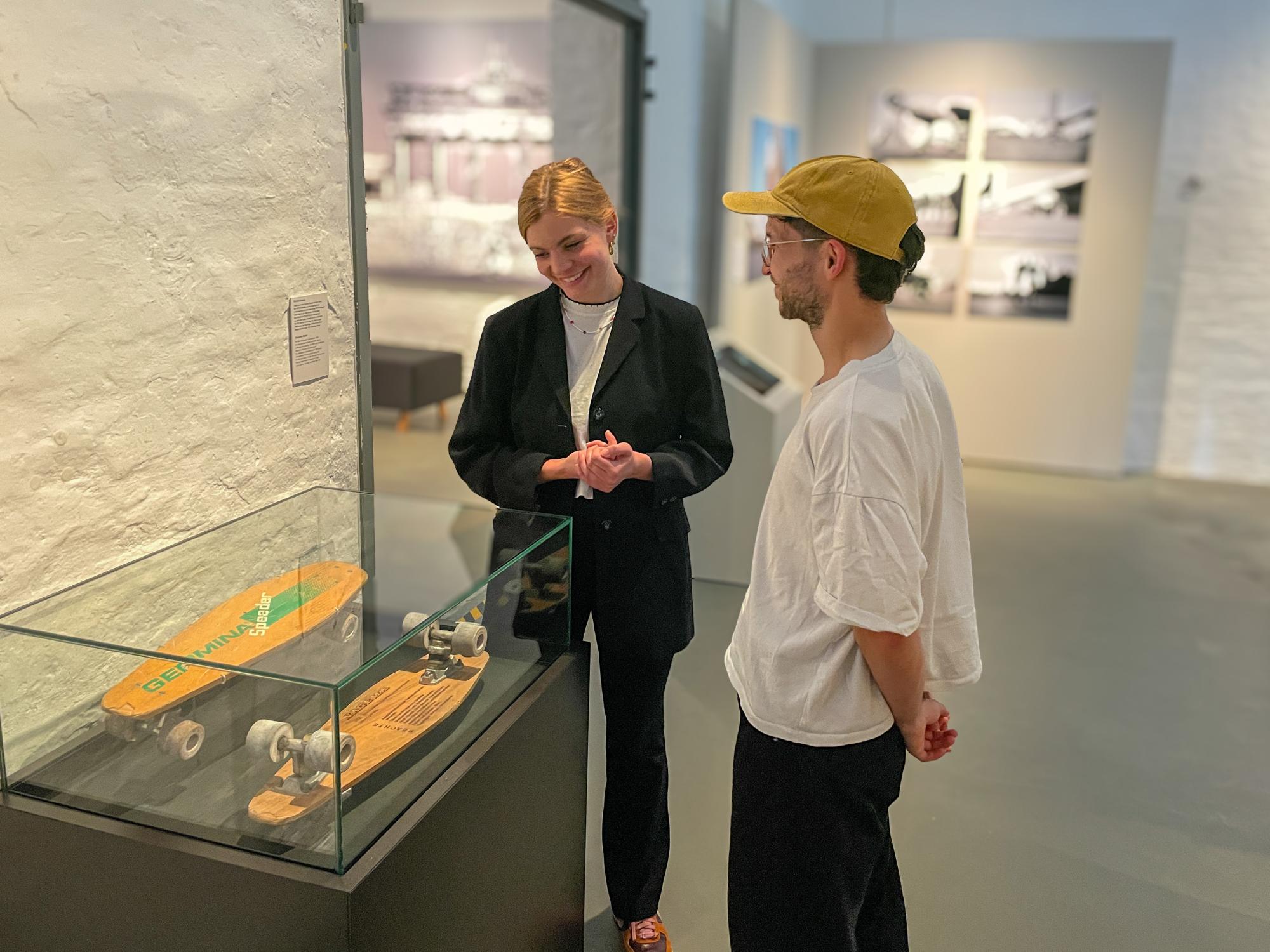 Ein Foto aus der Ausstellung: Eine Frau und ein Mann stehen vor einem Ausstellungskasten mit einem Skateboard darin.