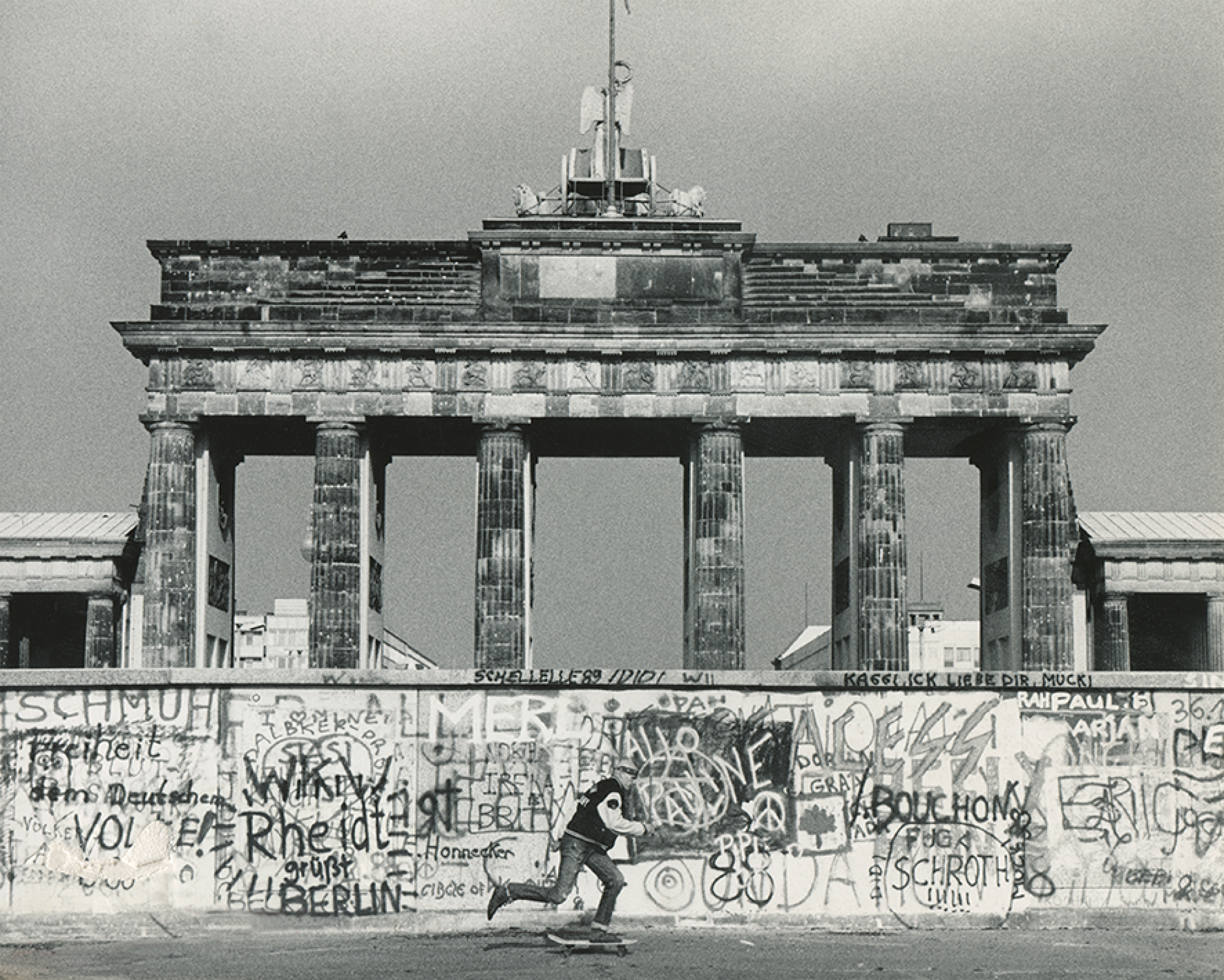 Ein Schwarzweiß-Foto des Brandenburger Tors mit der Mauer davor. Davor fährt ein Skateboarder.