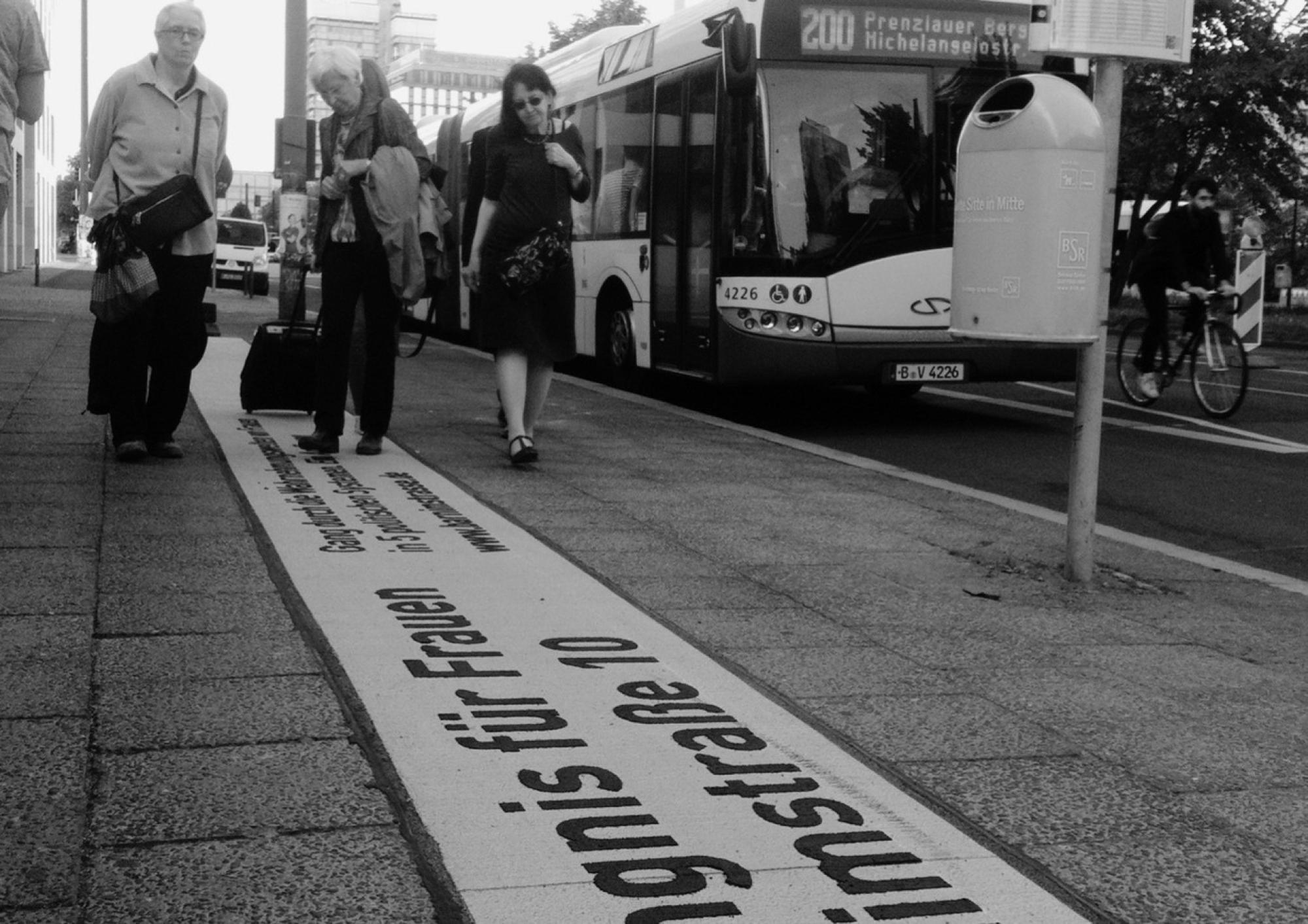 Ein Schwarzweiß-Foto von Menschen, die auf dem Schild "Gefändnis für Frauen Barnimstraße 10", das im Boden eingelassen ist, entlanggehen.