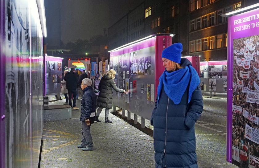 Menschen versammeln sich bei einer Open-Air-Veranstaltung auf dem Gelände der ehemaligen Stasi-Zentrale in Berlin; im Hintergrund das historische Gebäude, im Vordergrund bunte Infostände und Sitzgelegenheiten.