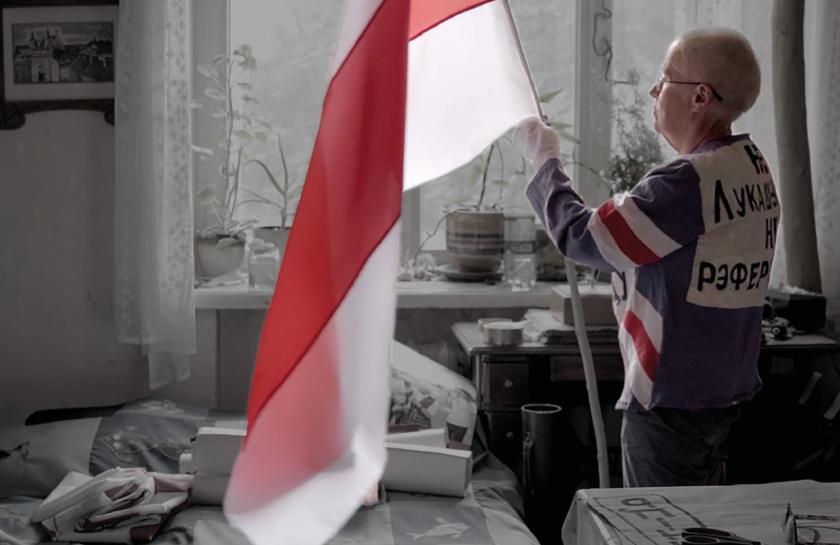 Ältere Frau mit kurzer Frisur hält die historische weiß-rot-weiße belarussische Flagge in einem privaten Zimmer; Text „Who, if not us?“ und „The Fight for Democracy in Belarus“ eingeblendet.