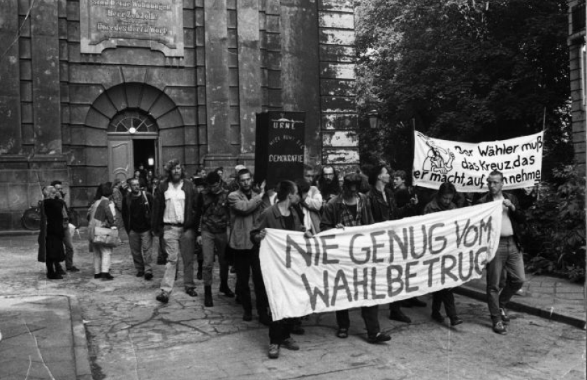 Schwarz-weiß-Fotografie einer Protestaktion in der DDR mit einer Gruppe von Menschen, die ein Banner mit der Aufschrift „Nie genug vom Wahlbetrug“ tragen. Im Hintergrund Transparente und ein historisches Gebäude.