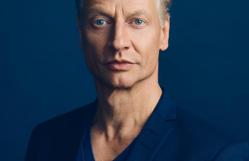 Porträt von Victor Schefé vor dunkelblauem Hintergrund, mit ernstem Gesichtsausdruck und blauen Augen, gekleidet in ein dunkelblaues Sakko und Shirt.