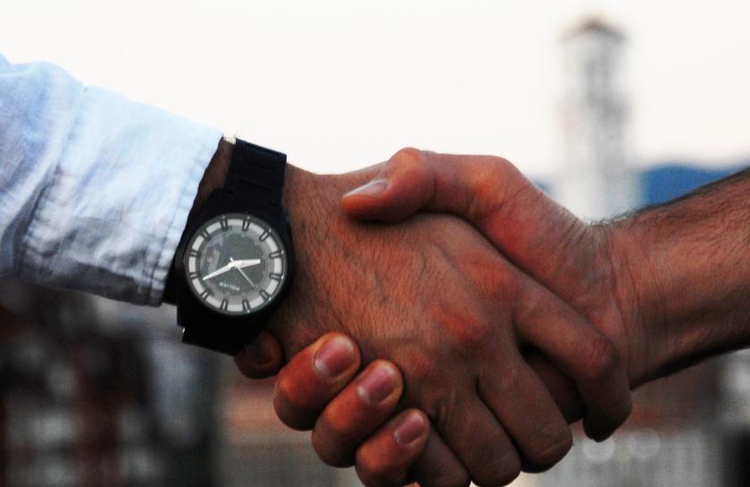 Ein Händedruck zwischen zwei weißen Männern. Ein Arm trägt eine schwarze Uhr und ein blaues Hemd.
