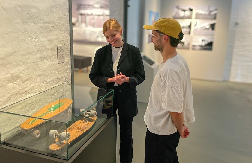 Ein Foto aus der Ausstellung: Eine Frau und ein Mann stehen vor einem Ausstellungskasten mit einem Skateboard darin.