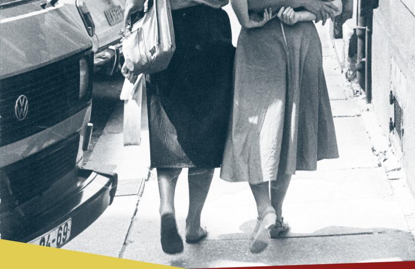 Ein Plakat für das Event "Lesbische Selbstbehauptung in der DDR." Eine Schwarzweiß-Fotografie von zwei Frauen, die Arm in Arm laufen.