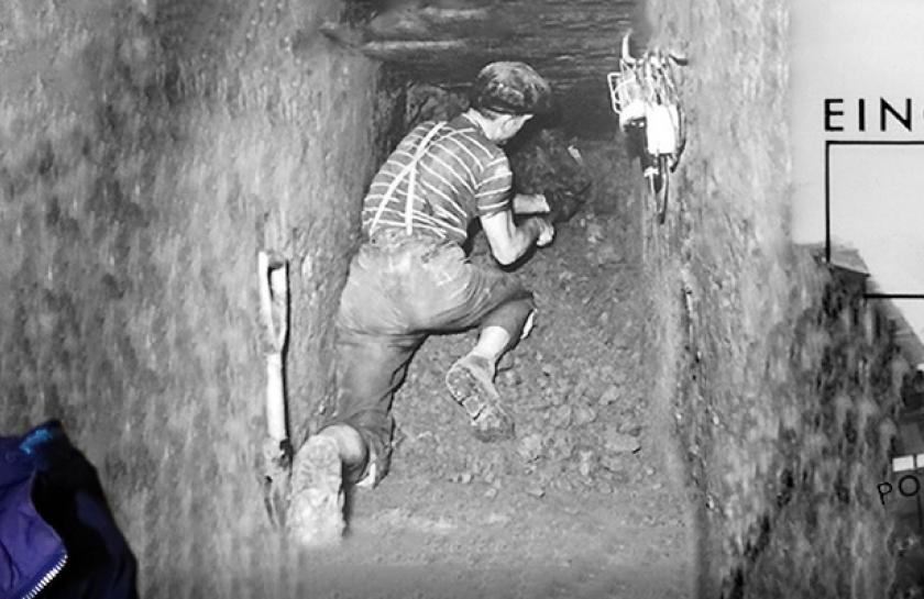 Ein Foto von Eveline Rudolph, einer Frau mit roten Haaren, vor einem schwarzweißen Foto eines Fluchttunnels.