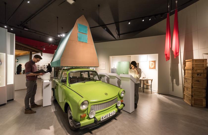 Ein Foto aus der Ausstellung: Eine Frau und ein Mann stehen neben einem alten Trabanten.